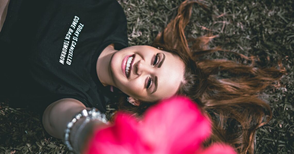 Overhead view of glad young female with makeup and colorful blooming flower looking at camera while lying on lawn